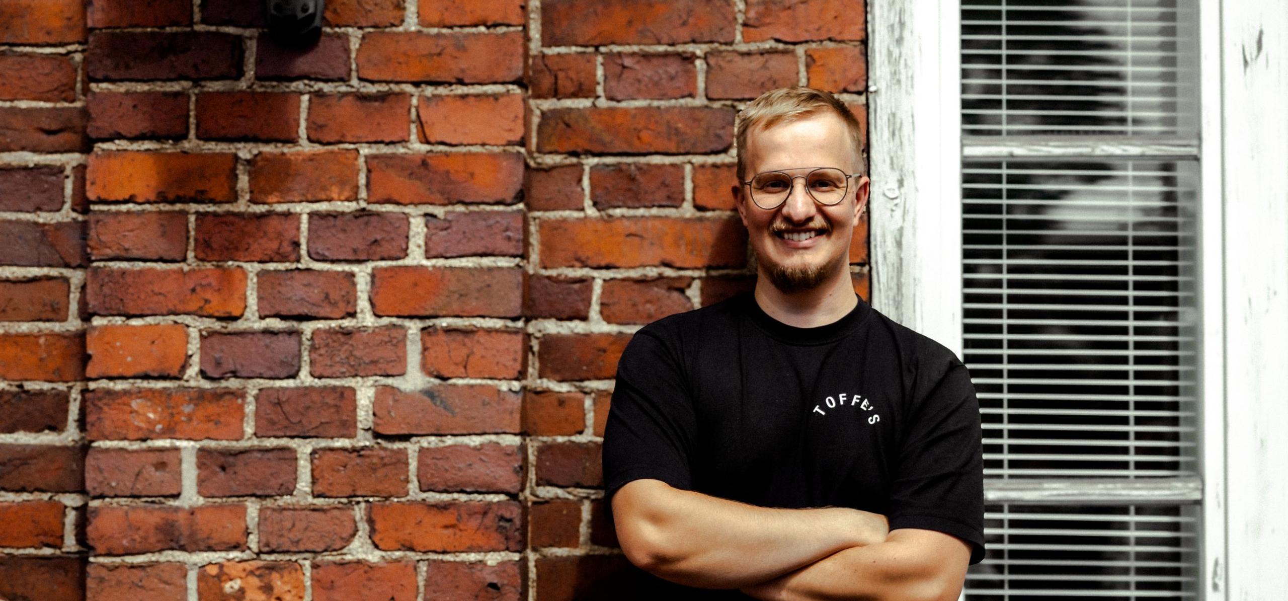 Toffe'sin yrittäjä Christoffer Holmström hymyilee tiiliseinän edessä. Ota yhteyttä lounas- ja catering-asioissa!